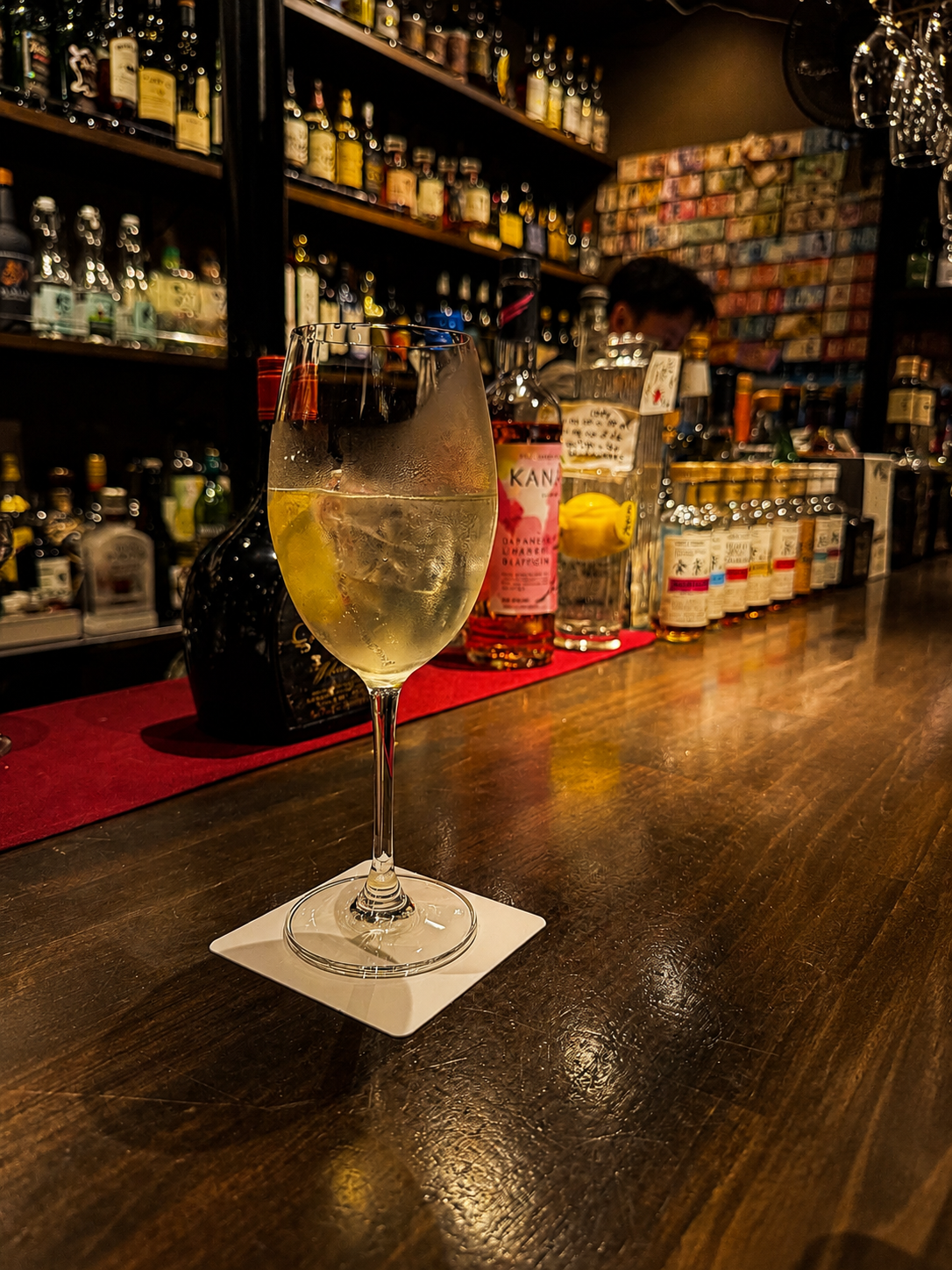 A glass of wine on the wooden counter at Bar Cordon Noir, backlit bottles behind, moody light