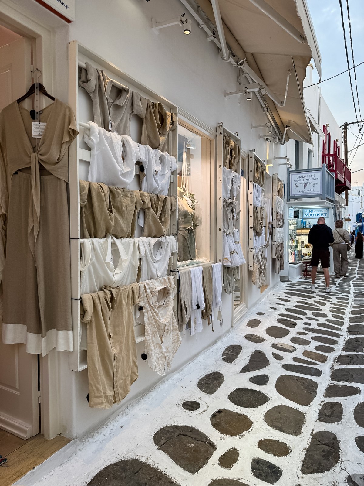 A narrow Chora lane with linen and cotton clothing hung on white walls outside a boutique, pebbled white-painted cobblestones underfoot