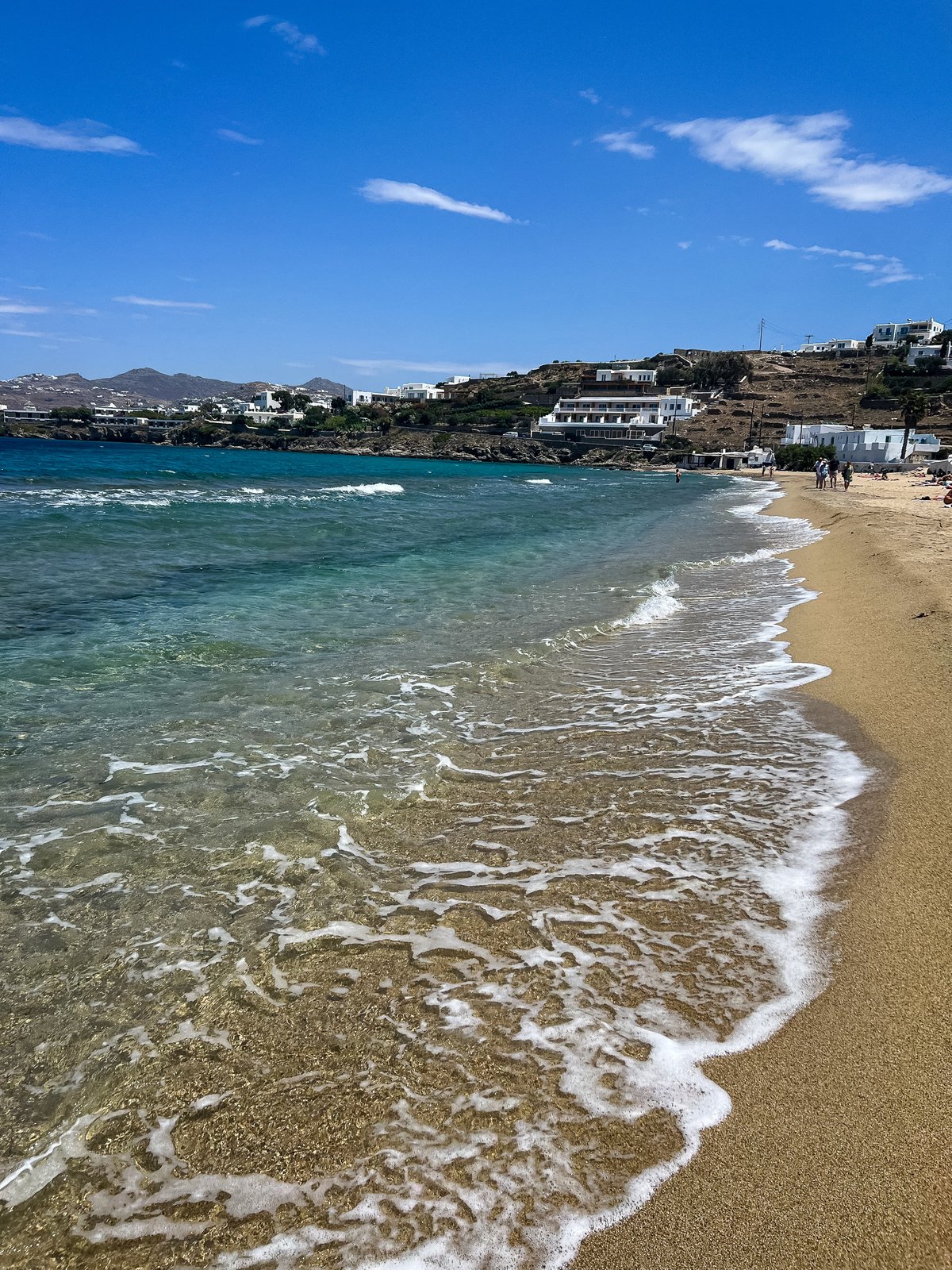 Wide view of Megali Ammos beach, soft waves rolling onto golden sand, turquoise-to-deep-blue water, whitewashed houses on the hillside beyond