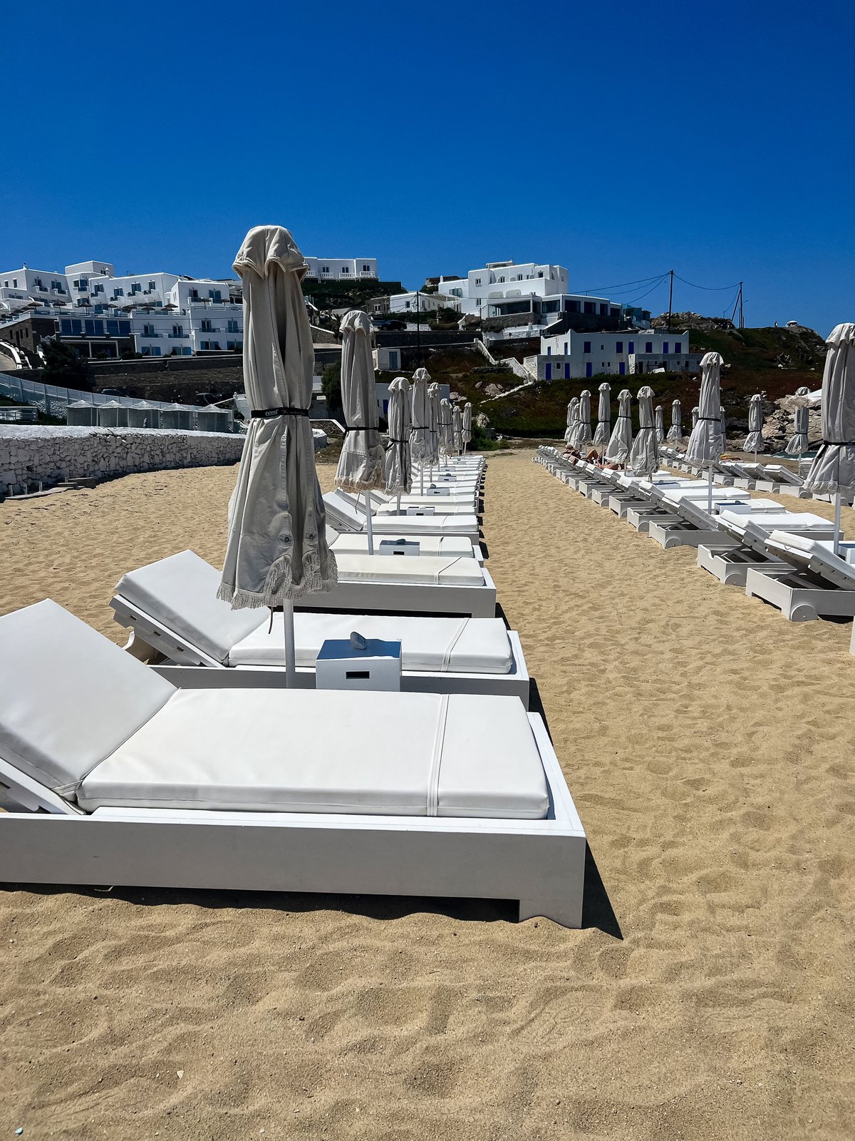 Neat rows of white sunbeds with folded cream umbrellas on the sand at Megali Ammos, whitewashed hotels stepping up the hill behind