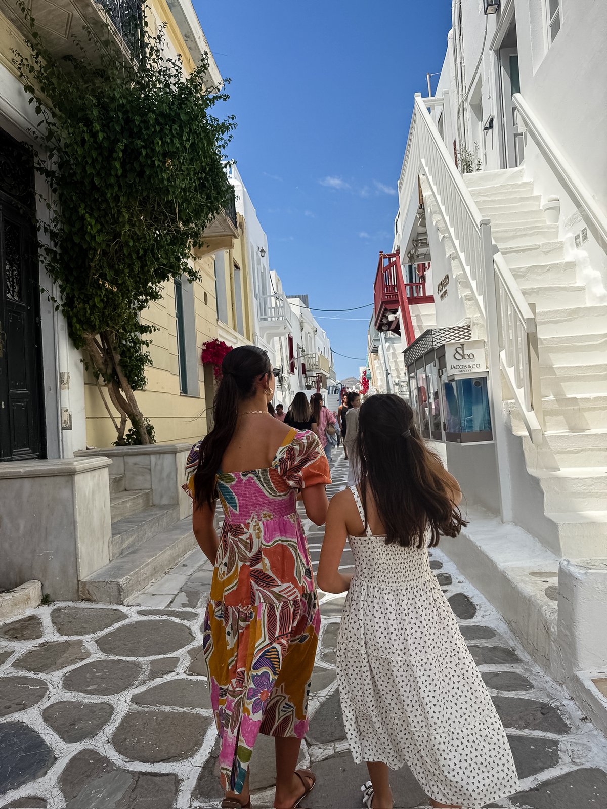 Two sisters in summer dresses walking hand in hand down a whitewashed, cobbled Chora lane lined with boutiques